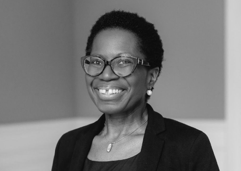 team-image1 A woman with short hair and glasses, wearing a blazer and necklace, smiles while standing indoors against a plain background.